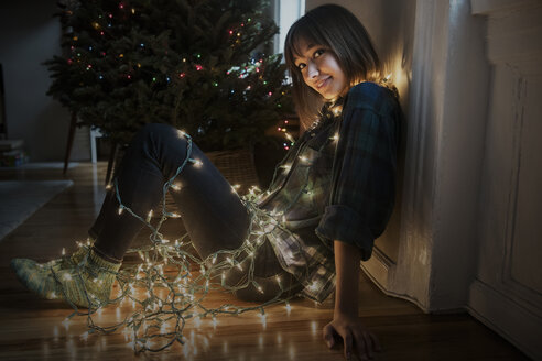 Mixed-Race-Frau sitzt auf dem Boden in Lichterketten eingewickelt in der Nähe von Weihnachtsbaum - BLEF01102