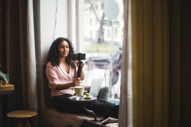 Confident young female influencer vlogging through smart phone on tripod sitting by window at creative office - MASF11915