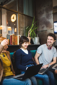 Lächelnde männliche und weibliche Kollegen, die eine weibliche Führungskraft betrachten, die mit einem Laptop auf einem Sofa im Kreativbüro sitzt - MASF11924