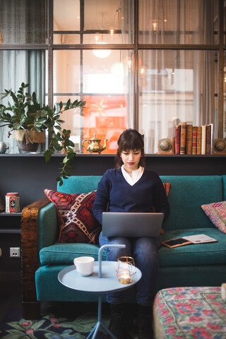 Selbstbewusste Frau in voller Länge mit Laptop auf dem Sofa im Büro sitzend, lizenzfreies Stockfoto