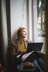 Nachdenkliche reife Frau, die mit ihrem Laptop auf der Fensterbank im Büro sitzt und wegschaut - MASF11935