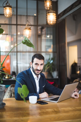 Porträt eines selbstbewussten männlichen Berufstätigen, der einen Laptop benutzt, während er am Tisch im Büro sitzt, lizenzfreies Stockfoto