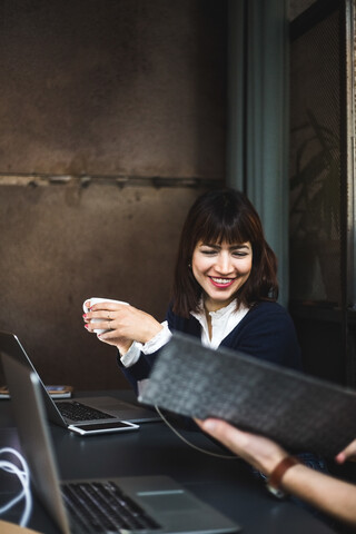 Lächelnde Unternehmerin, die eine Kaffeetasse hält und mit einem Kollegen über ein Dokument am Schreibtisch im Büro diskutiert, lizenzfreies Stockfoto