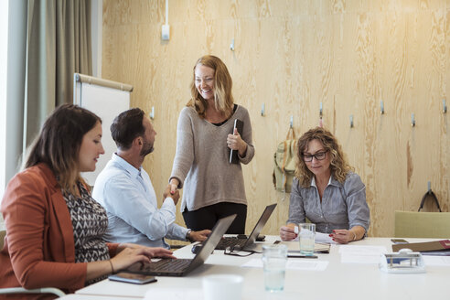 Lächelnde Geschäftsfrau, die einem Geschäftsmann die Hand schüttelt, während weibliche Kollegen am Konferenztisch arbeiten - MASF11982