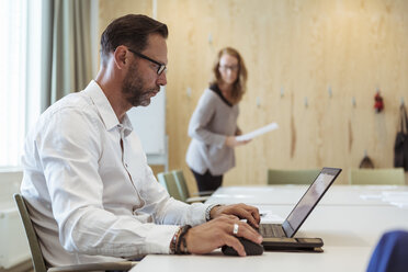 Seriöser Geschäftsmann mit Laptop am Konferenztisch mit einer Kollegin im Hintergrund - MASF11990