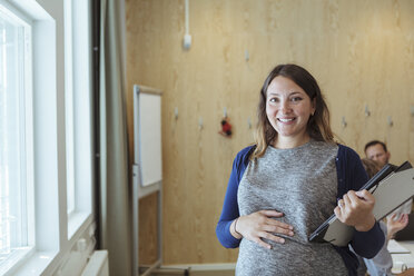 Portrait of smiling pregnant businesswoman with file and laptop standing in office - MASF12001