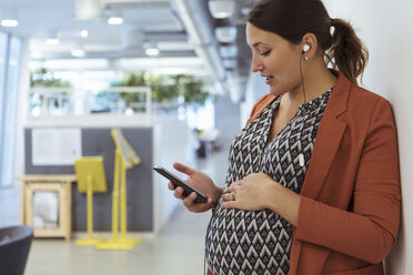 Schwangere Geschäftsfrau hört Musik auf Smartphone mit Händen auf dem Bauch im Büro - MASF12017