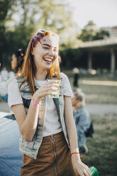 Cheerful woman having drink while standing at lawn in music festival - MASF12078