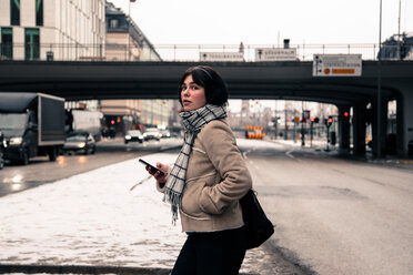 Side view of young woman holding mobile phone while crossing road in city during winter - MASF12127