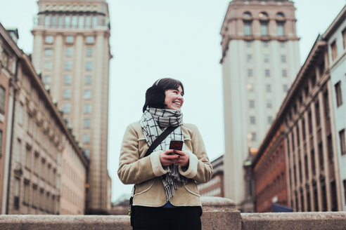 Fröhliche junge Frau, die wegschaut, während sie ihr Handy gegen Gebäude in der Stadt im Winter hält - MASF12128