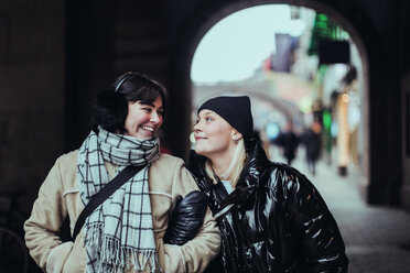 Smiling young female friends looking each other face to face while standing on street in city during winter - MASF12129