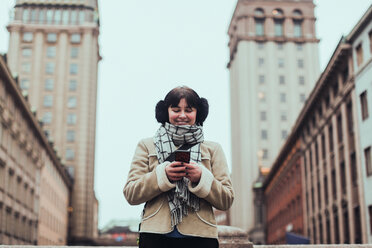 Lächelnde junge Frau in warmer Kleidung bei der Benutzung eines Mobiltelefons vor einem Gebäude in der Stadt - MASF12130