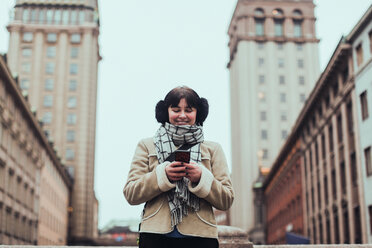 Smiling young woman wearing warm clothing while using mobile phone against buildings in city - MASF12130