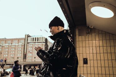 Side view of young woman wearing black shiny jacket while standing at subway's entrance in city during winter - MASF12144