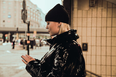 Side view of young woman using mobile phone while wearing black shiny jacket at subway's entrance in city during winter - MASF12145