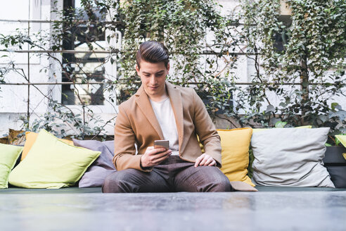 Junger Mann sitzt auf einer Couch in einem Café und benutzt ein Mobiltelefon - FBAF00435