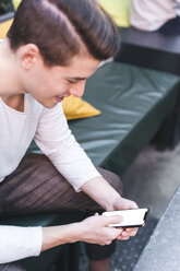 Smiling young man sitting on couch in a cafe using cell phone - FBAF00439