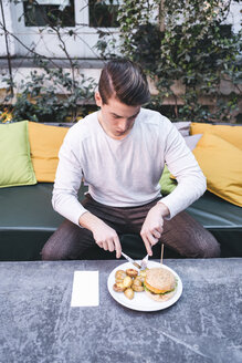 Junger Mann sitzt auf einer Couch in einem Restaurant und isst einen veganen Burger zum Mittagessen - FBAF00444