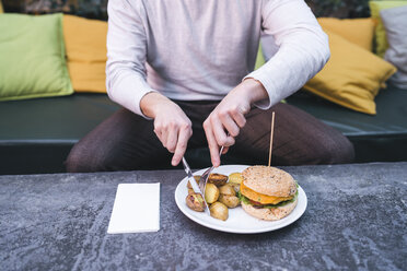 Junger Mann sitzt auf einer Couch in einem Restaurant und isst einen veganen Burger mit Kartoffeln zum Mittagessen - FBAF00445