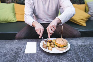 Young man sitting on couch in a restaurant having a vegan burger with potatoes for lunch - FBAF00445