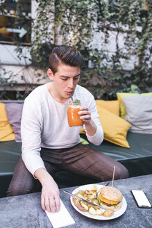 Junger Mann sitzt auf einer Couch in einem Restaurant und isst einen veganen Burger und ein gesundes Getränk zum Mittagessen - FBAF00446