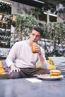 Junger Mann sitzt auf einer Couch in einem Restaurant und isst einen veganen Burger und ein gesundes Getränk zum Mittagessen - FBAF00447