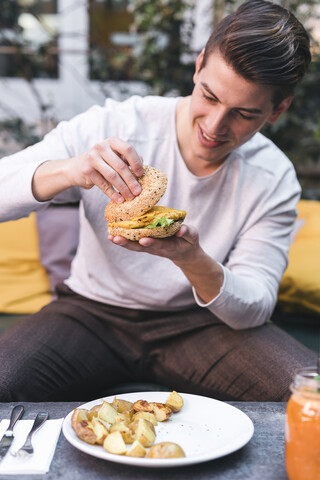 Junger Mann sitzt auf einer Couch in einem Restaurant und isst einen veganen Burger zum Mittagessen, lizenzfreies Stockfoto