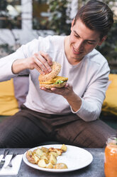 Junger Mann sitzt auf einer Couch in einem Restaurant und isst einen veganen Burger zum Mittagessen - FBAF00450