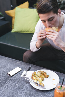 Junger Mann sitzt auf einer Couch in einem Restaurant und isst einen veganen Burger zum Mittagessen - FBAF00451