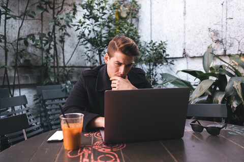 Junger Mann sitzt am Tisch und benutzt einen Laptop, lizenzfreies Stockfoto