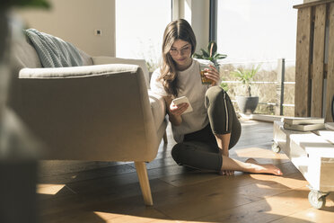 Junge Frau sitzt zu Hause mit Getränk und Handy auf dem Boden - UUF17267