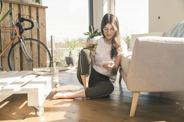 Young woman sitting on the floor at home with drink and cell phone - UUF17268