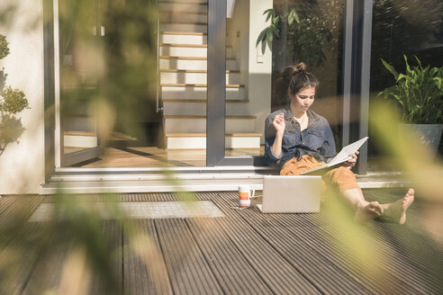 Junge Frau sitzt auf der Terrasse zu Hause und arbeitet mit Buch und Laptop - UUF17300