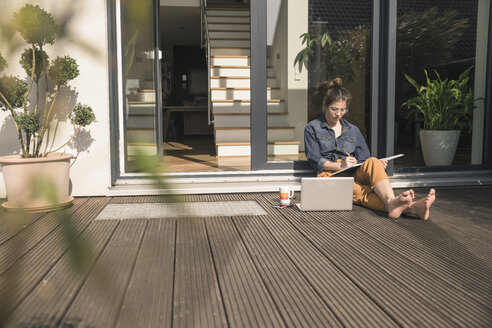 Junge Frau sitzt auf der Terrasse zu Hause und arbeitet mit Buch und Laptop - UUF17301