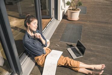 Junge Frau mit geschlossenen Augen sitzt auf der Terrasse zu Hause mit Laptop und Buch - UUF17305