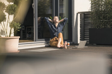Junge Frau mit geschlossenen Augen sitzt auf der Terrasse zu Hause mit Laptop - UUF17306