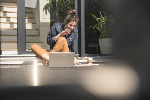 Junge Frau sitzt auf der Terrasse zu Hause und benutzt Handy und Laptop - UUF17307