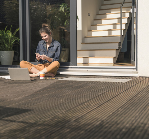 Happy young woman sitting on terrace at home using cell phone and laptop stock photo