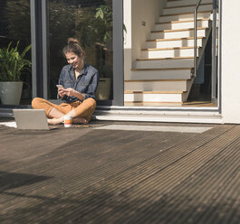 Glückliche junge Frau sitzt auf der Terrasse zu Hause und benutzt Handy und Laptop - UUF17308