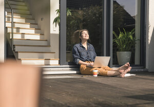 Junge Frau mit geschlossenen Augen sitzt auf der Terrasse zu Hause mit Laptop - UUF17309