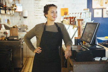 Confident young female owner standing with hand on hip by cash register at checkout in cafe - MASF12284