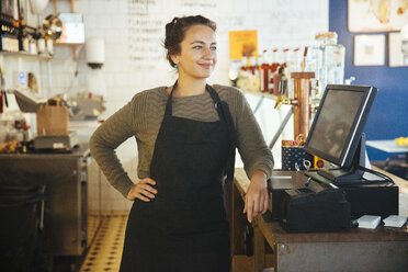 Selbstbewusste junge Besitzerin steht mit der Hand auf der Hüfte an der Kasse eines Cafés - MASF12284