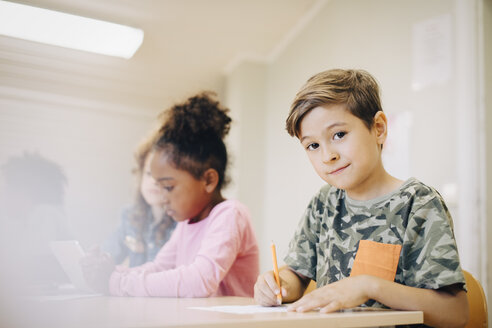 Porträt eines Jungen, der mit einem Freund an einem Schreibtisch im Klassenzimmer sitzt und auf Papier schreibt - MASF12300