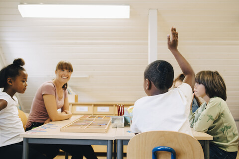 Lehrer sieht Junge an, der die Hand hebt, während er im Klassenzimmer antwortet, lizenzfreies Stockfoto