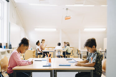 Boy and girl writing at desk while friends learning with student in background - MASF12319