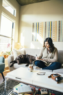 Mixed Race woman on sofa with dog writing in journal - BLEF01243