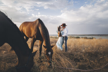 Weidende Pferde in der Nähe einer kaukasischen Frau, die einen Cowboy umarmt - BLEF01274