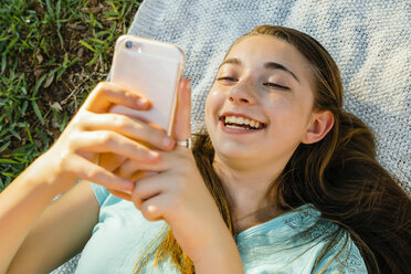Lachendes kaukasisches Mädchen, das auf einer Decke liegt und eine SMS auf einem Handy schreibt - BLEF01421