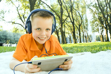 Lächelnder kaukasischer Junge, der auf einer Decke im Park liegt und einem digitalen Tablet zuhört - BLEF01427