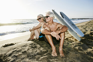 Older Caucasian couple sitting on beach with surfboards - BLEF01755